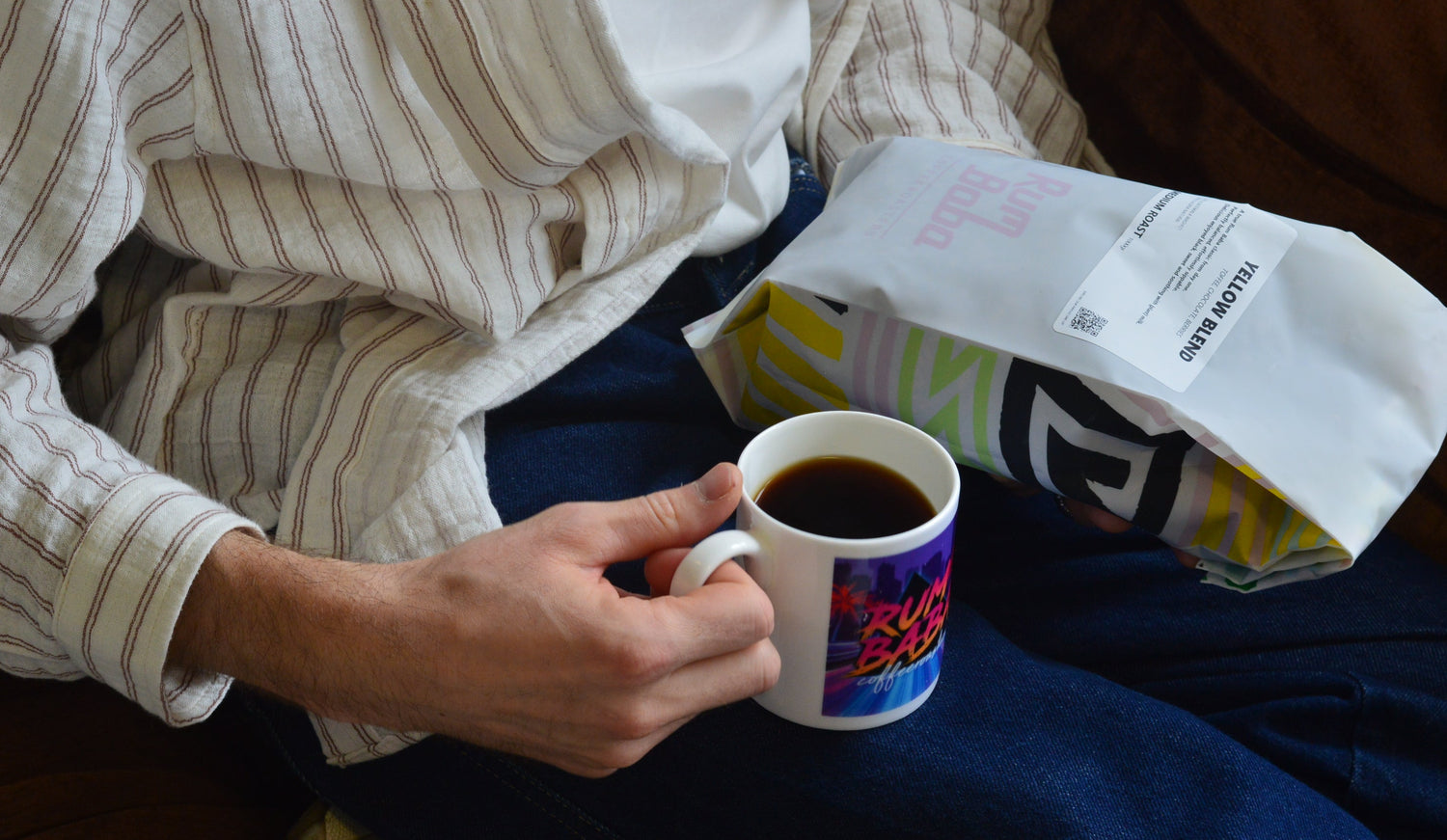 Person holding a retro coffee mug with a colorful design, sitting on a couch with a coffee bag beside them.